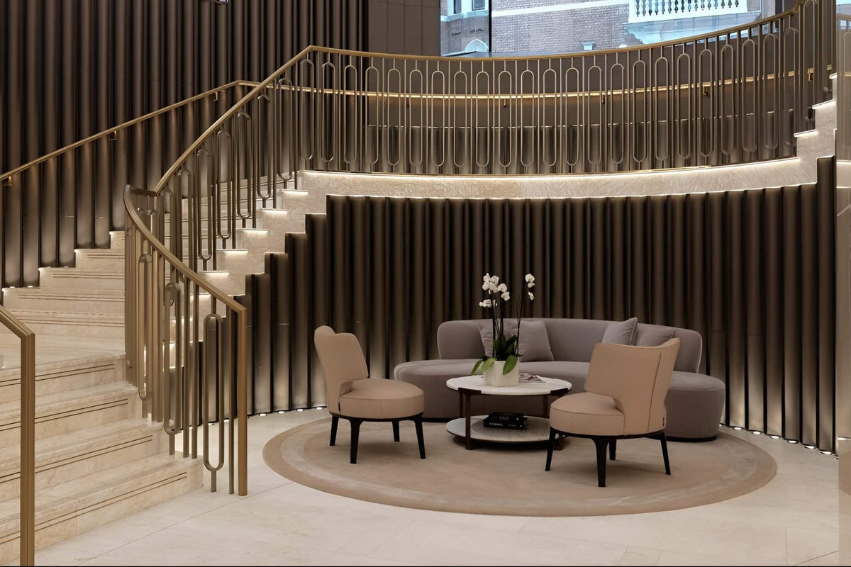 A luxury modern interior featuring a sweeping curved staircase with gold-toned metal railings and light stone steps, overlooking a circular seating area with a gray sofa and two beige armchairs on a round rug.