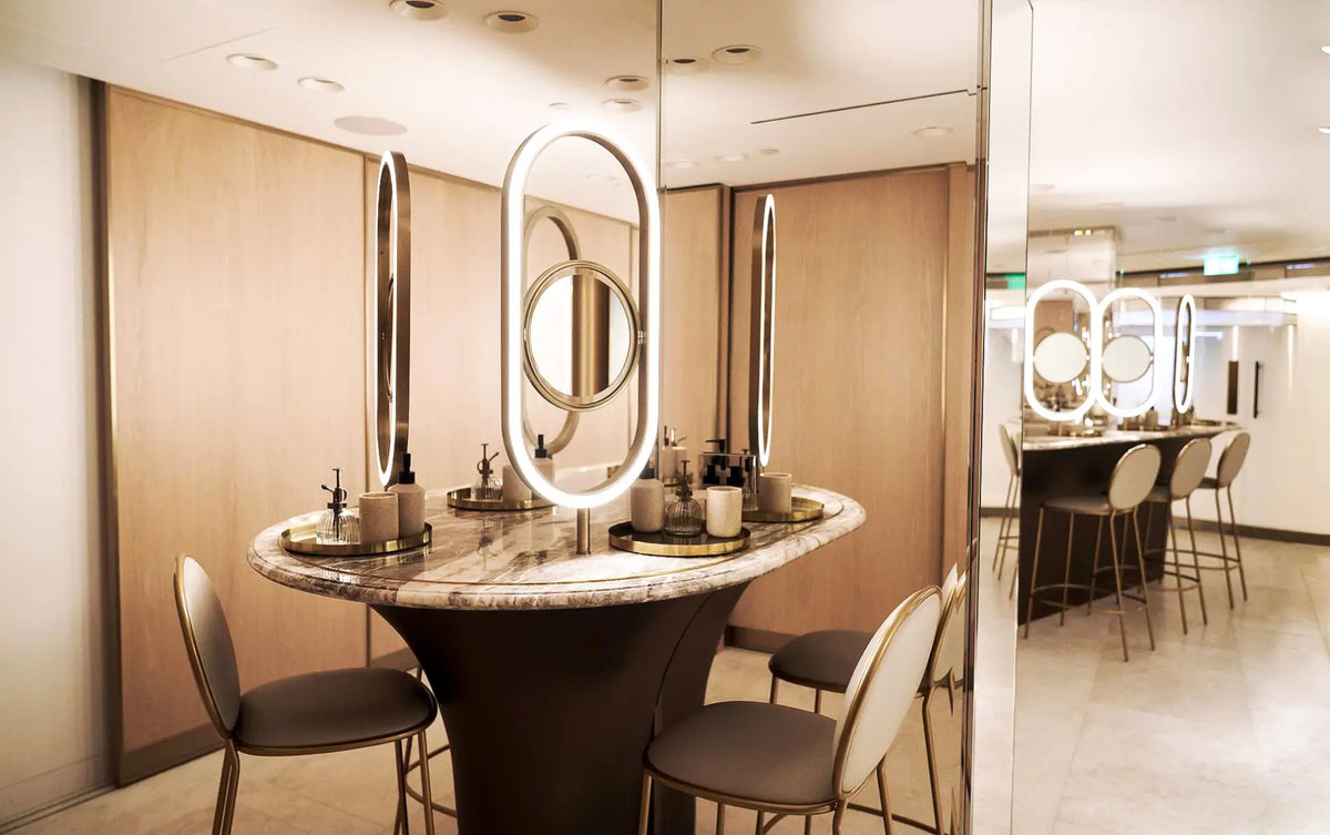 A modern vanity station with a round marble-top console table, dark pedestal base, and multiple illuminated oval mirrors, surrounded by upholstered chairs in a warm interior setting.
