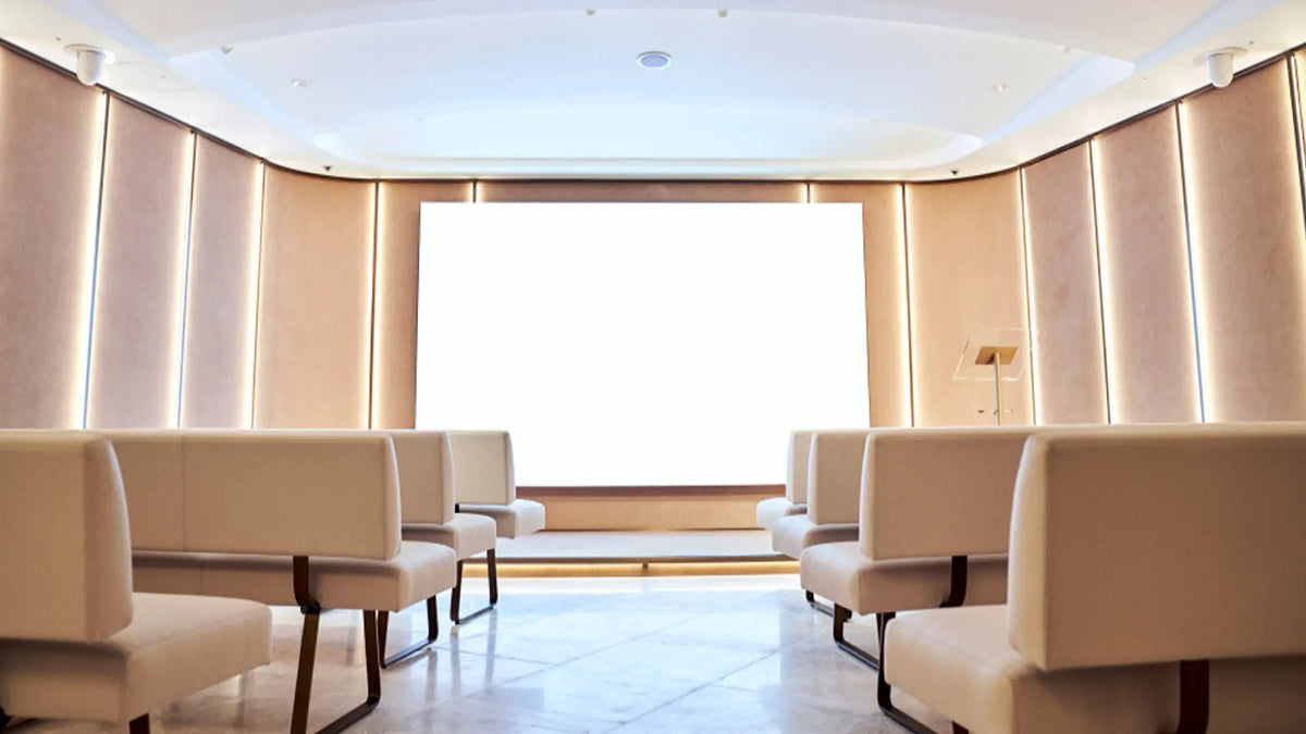 A modern, minimalist event hall or auditorium with rows of cream-colored upholstered chairs facing a large, bright white projection screen. The room features warm beige and light wood-paneled walls with integrated LED lighting, a polished light gray floor, and a white ceiling.