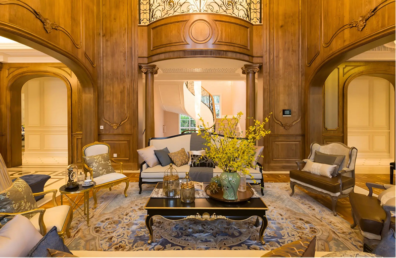 A luxury high-ceilinged living room with ornate wood-paneled walls, arched entryways, and a central staircase. The room is furnished with classic sofas, armchairs, a dark wood coffee table, and a large patterned area rug.