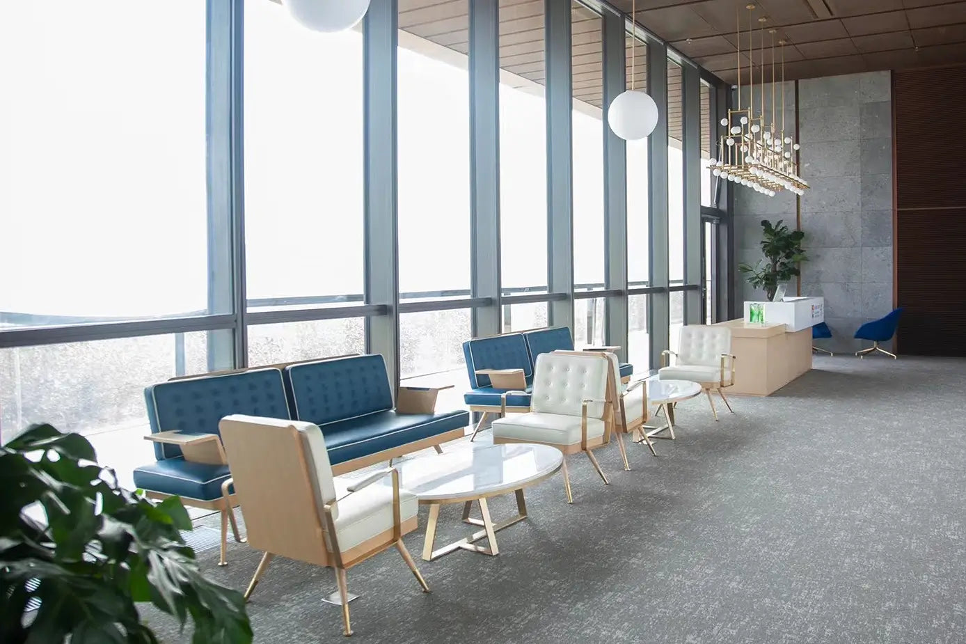 A modern office lounge area with blue and cream upholstered seating, round white coffee tables, tall floor-to-ceiling windows, and a reception desk in the background.