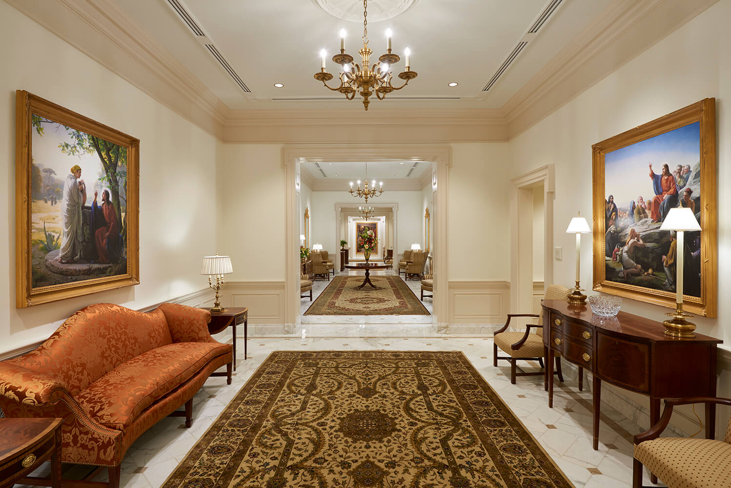A grand, elegant church interior hallway with cream walls, a large patterned oriental rug, a rust-orange velvet sofa on the left, a dark wood console table with lamps on the right, and two large framed classical paintings on the walls.