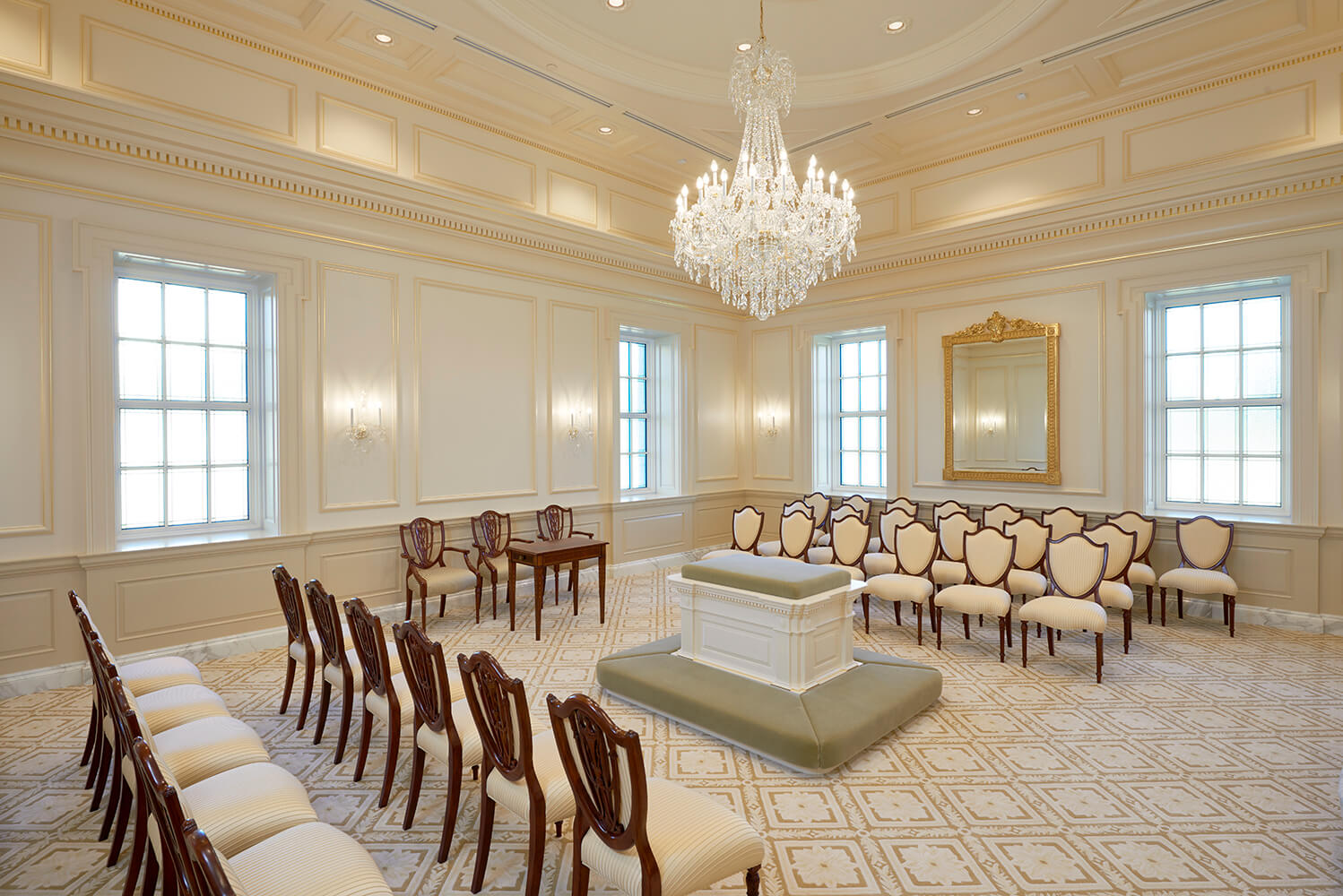 A grand church with rows of cream and dark wood chairs arranged around a central podium, featuring a large crystal chandelier, patterned carpet, and tall windows.
