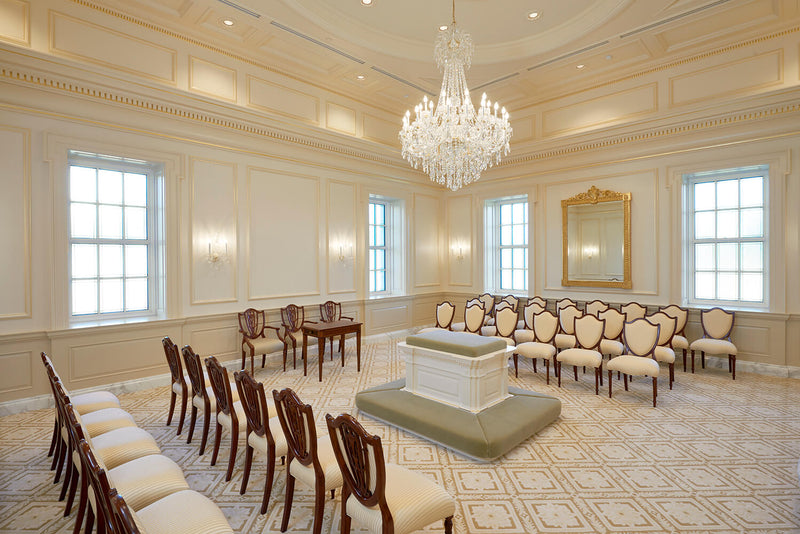 A grand church with rows of cream and dark wood chairs arranged around a central podium, featuring a large crystal chandelier, patterned carpet, and tall windows.