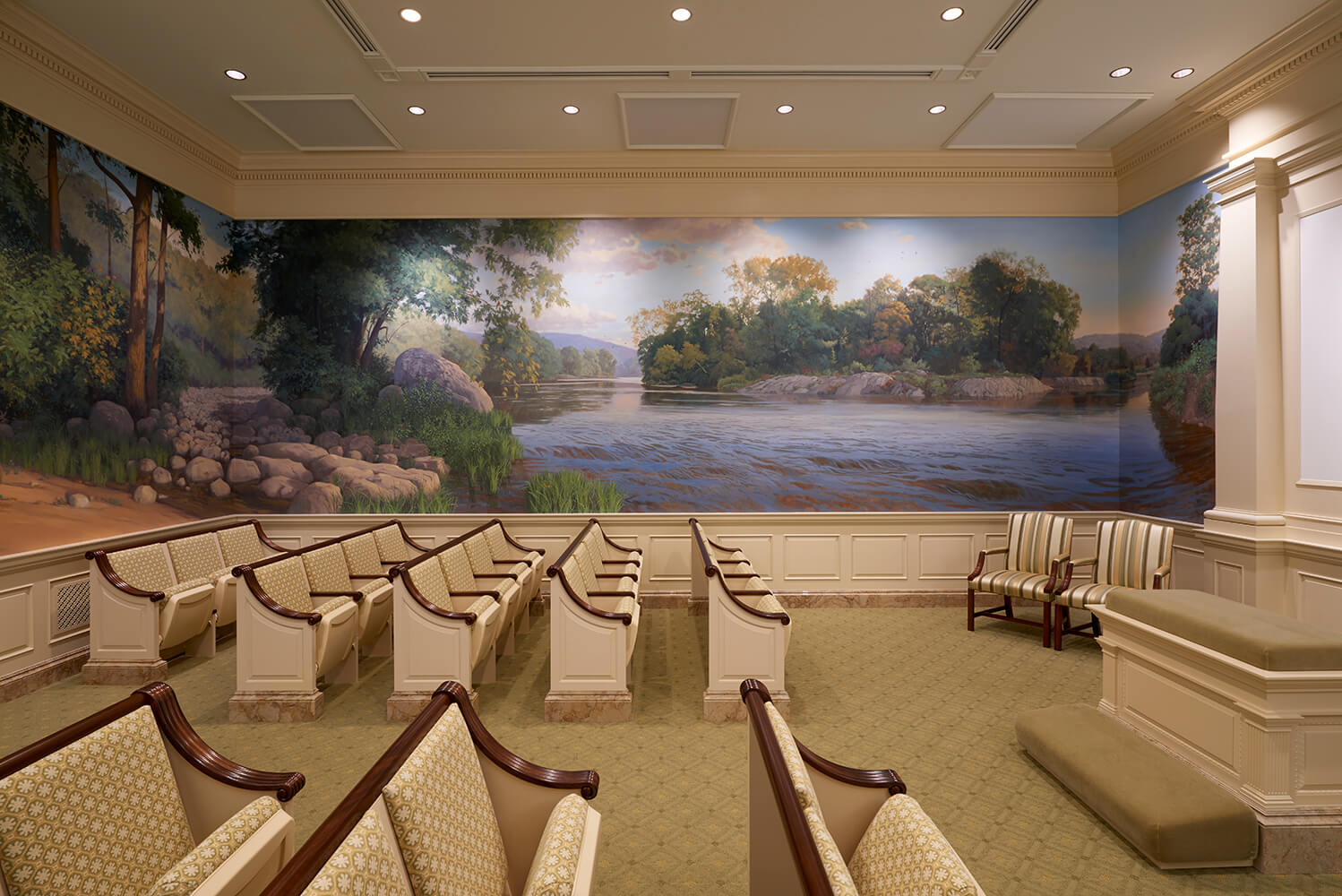 A large church interior room with rows of upholstered chairs facing a massive scenic mural depicting a river landscape with trees, rocks, and distant hills. The room has cream walls, recessed ceiling lights, and a carpeted floor.