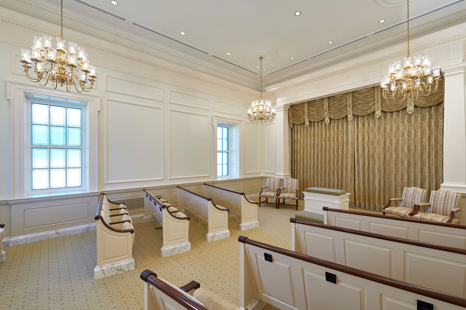 A spacious, elegant church interior room with cream walls, decorative molding, two large chandeliers, patterned gold drapery, and rows of wooden pews with cream upholstery.
