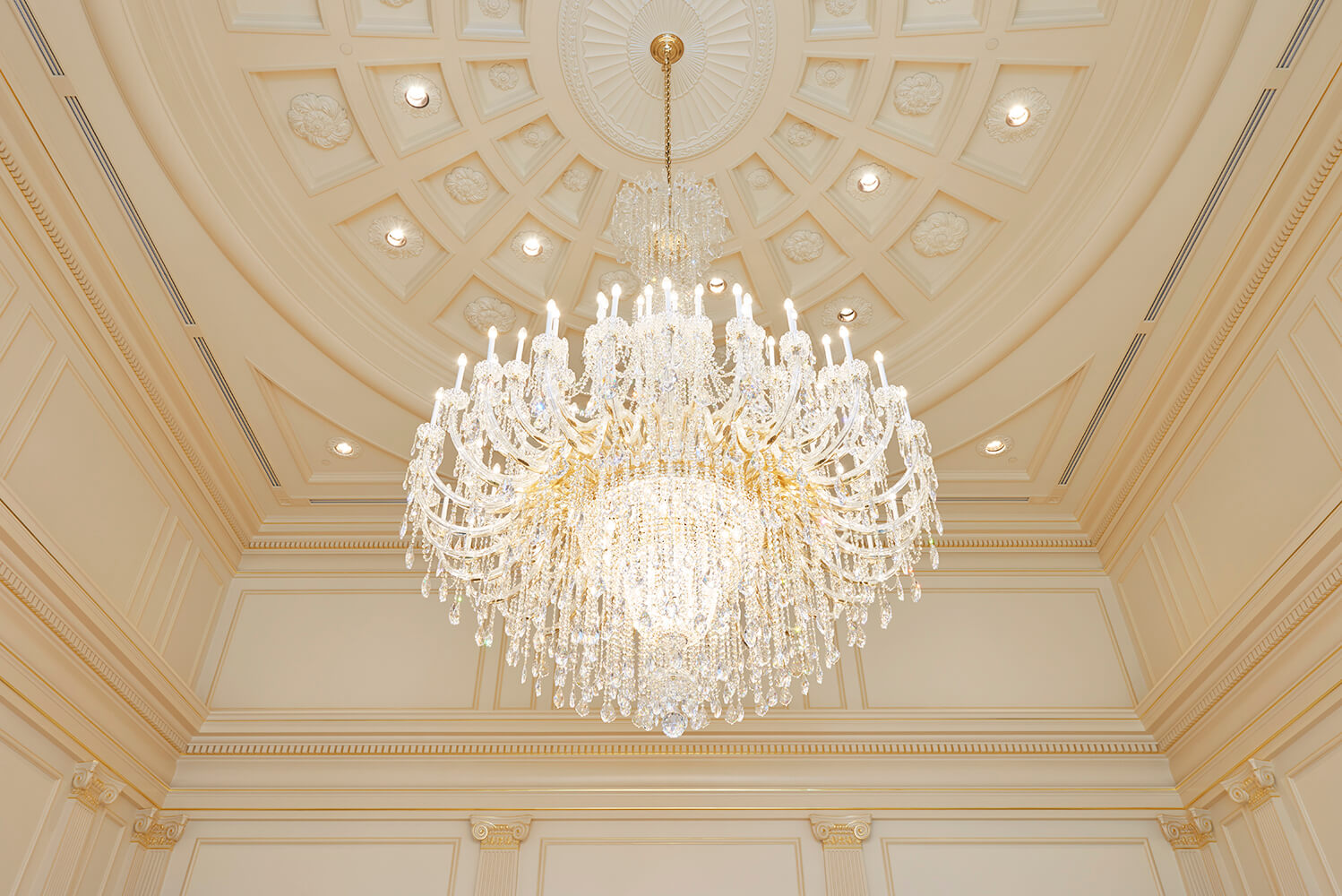 A large ornate crystal chandelier with gold-toned metal details hangs from a high, decorated cream-colored coffered ceiling with recessed lights.