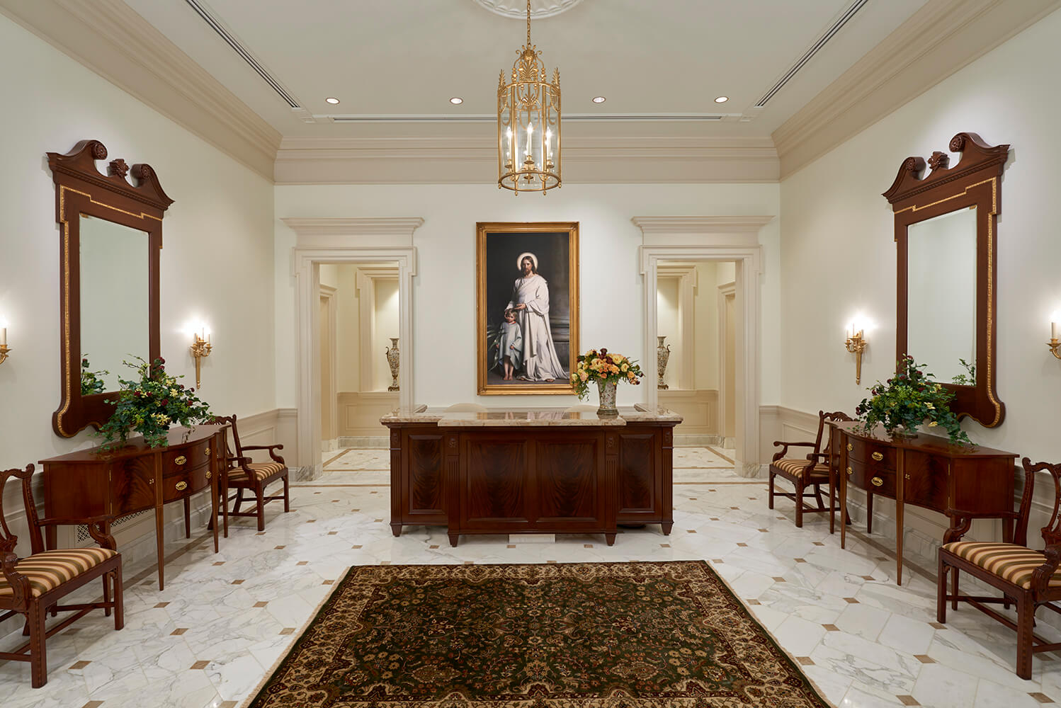An elegant church interior hall with a large framed portrait on the back wall, a central wooden console table, matching side tables and chairs, tall decorative mirrors, sconces, and a patterned area rug on a light marble floor.