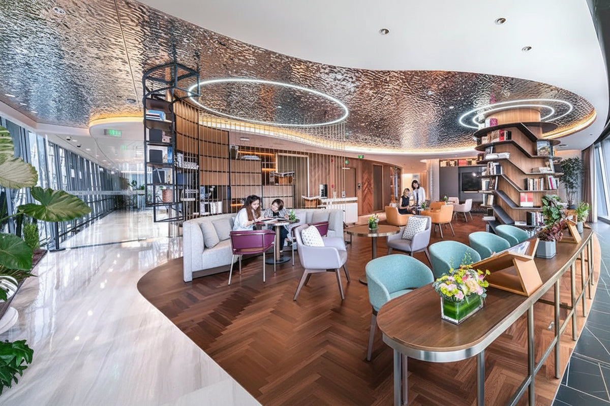 A modern, spacious hotel lobby or lounge interior with a curved ceiling, textured metallic pattern, wood flooring, white and blue lounge chairs, shelving, and large windows.