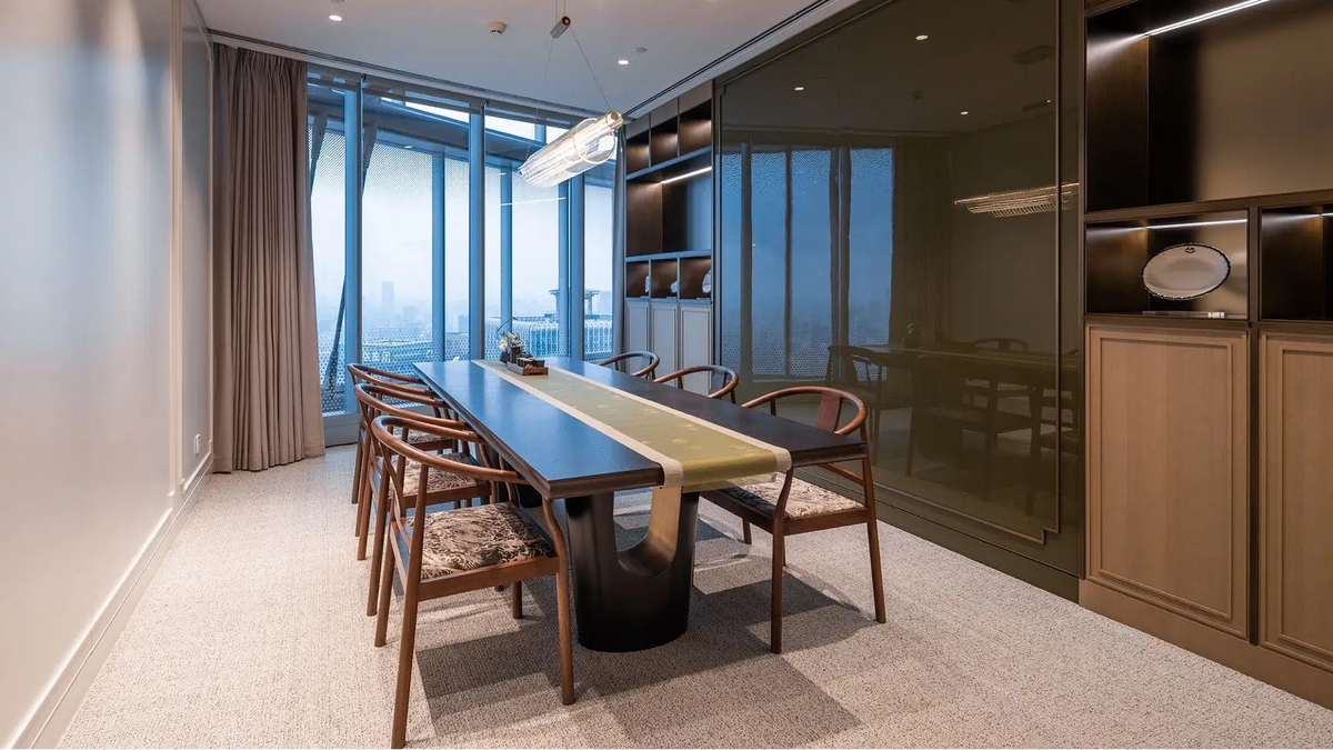 A modern dining room featuring a long rectangular wooden table with a black pedestal base, surrounded by six wooden chairs with patterned seat cushions. The room has a large glass window with beige curtains, a dark green glass wall, and built-in wooden cabinets with integrated lighting on the right side.