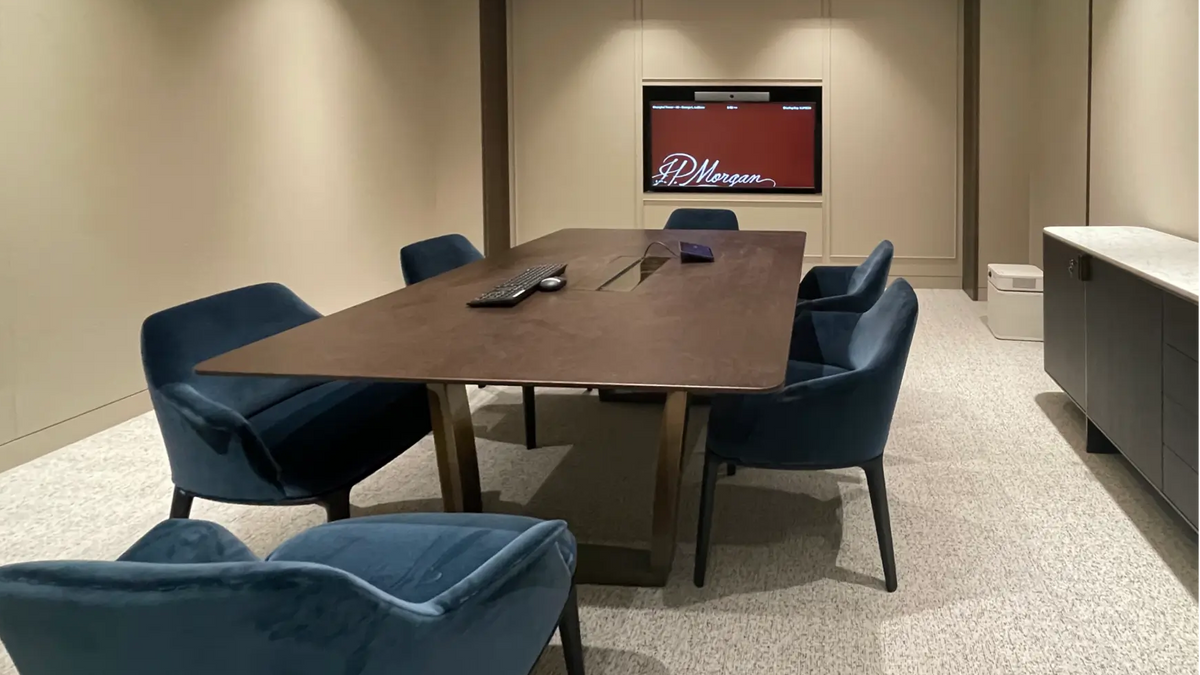 A modern conference room setup featuring a rectangular dark wood table surrounded by six blue velvet armchairs, set against a beige wall with a built-in television.