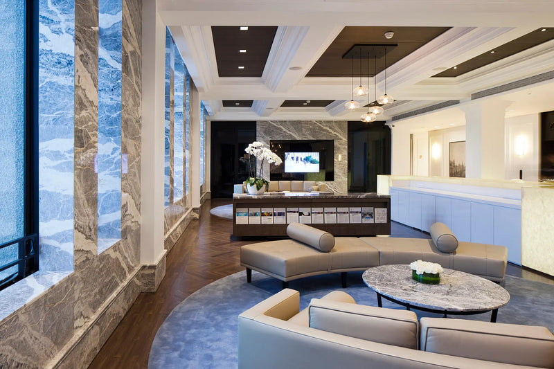 A luxury modern lobby lounge with curved light gray sofas, a round marble coffee table, a reception desk, and large windows with blue-tinted marble frames.