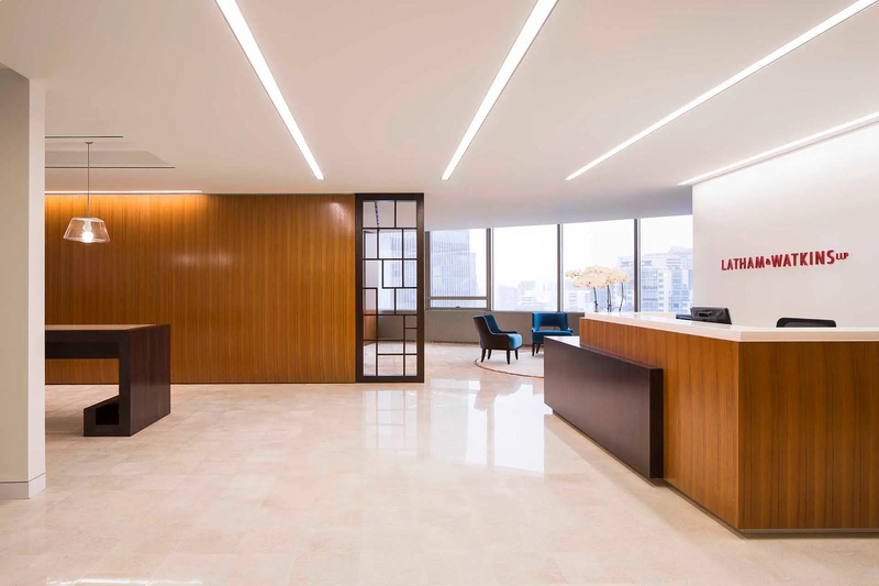 A modern office lobby with a wood-paneled reception desk, a dark wood desk on the left, and large windows in the background. The wall displays the red text 'LATHAM+WATKINS'.
