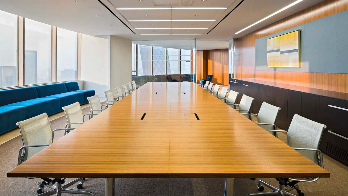 A modern conference room with a long rectangular wooden table, multiple white office chairs, large windows, a blue upholstered bench, wood-paneled walls, and a framed abstract artwork.