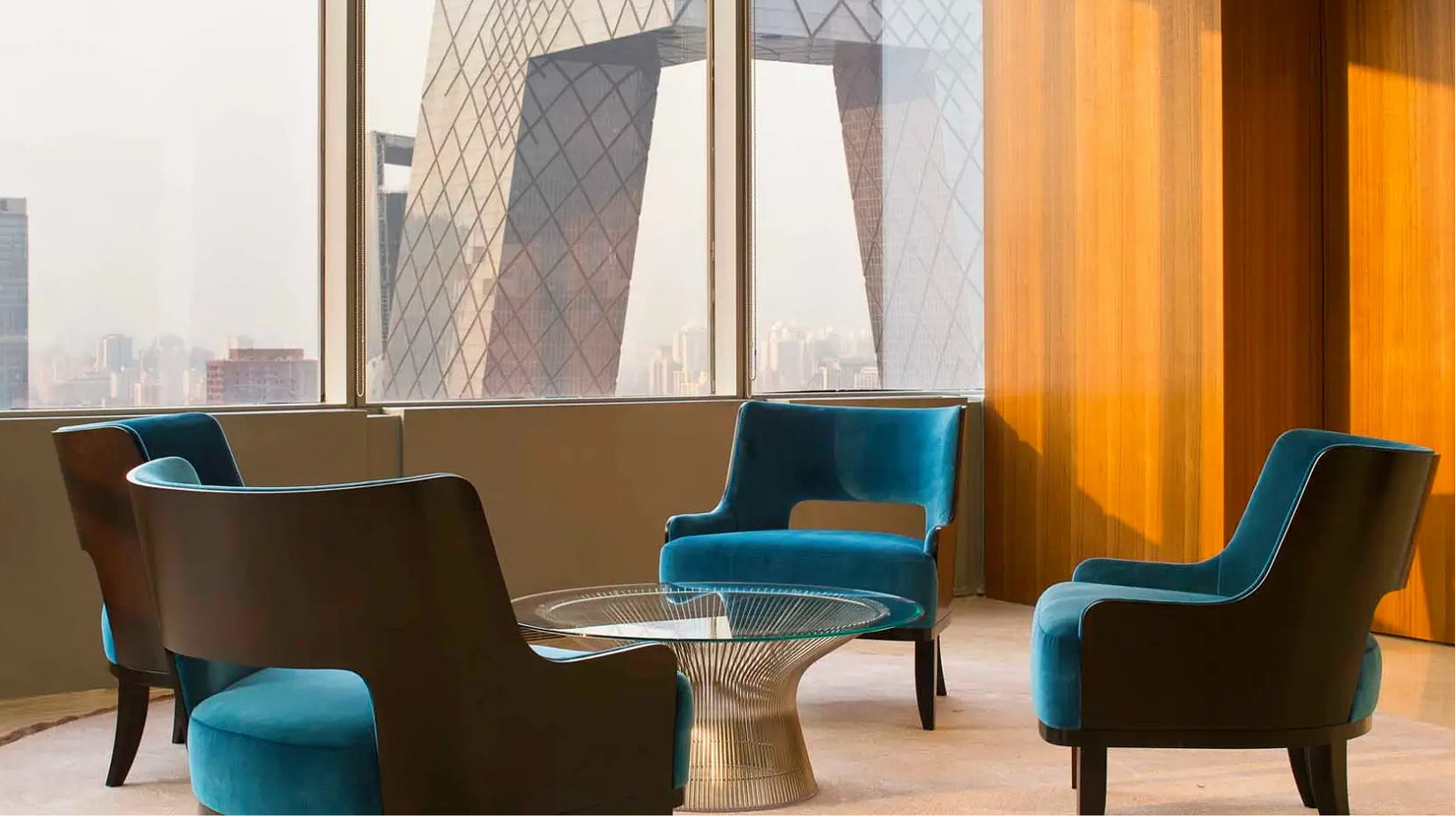 A modern interior lounge area with four upholstered armchairs around a small round glass-top table. The chairs are in teal blue and dark brown, set in a room with warm wood-paneled walls and large windows overlooking a city skyline.