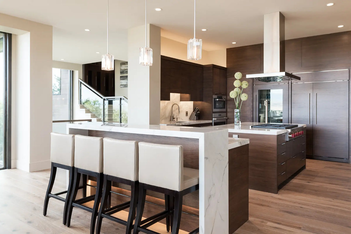 A modern kitchen island with a white marble-look countertop and base, paired with four upholstered bar stools featuring cream seats and black legs. The background showcases dark wood cabinetry, stainless steel appliances, and a large white range hood, all set in a bright, contemporary kitchen with wood floors.