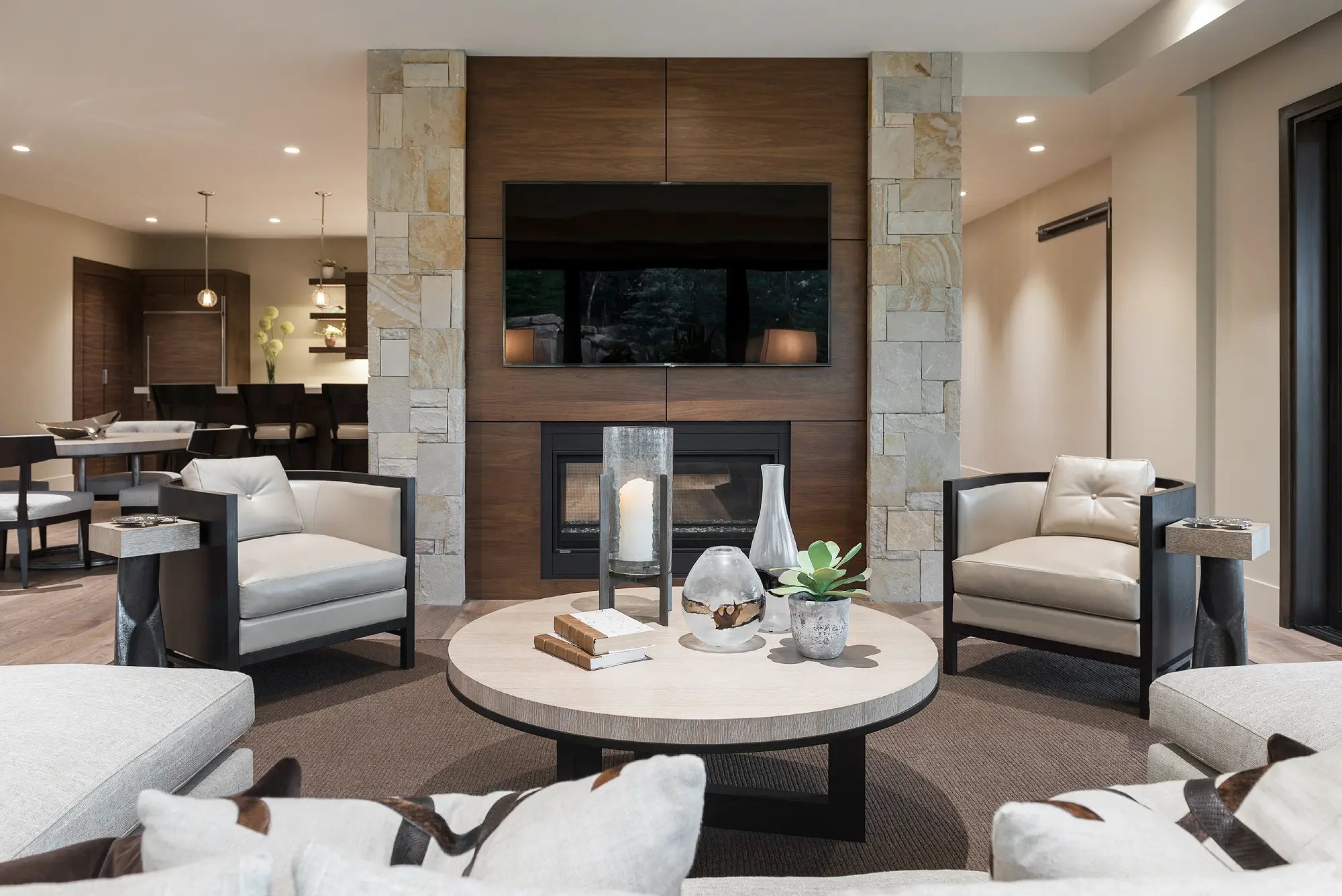 A modern living room interior featuring a round light-wood coffee table with decorative glassware and books in the center, flanked by two light gray armchairs with dark wood frames. The room has a large fireplace with a dark wood surround and stone columns, a mounted TV, and a dining area in the background.