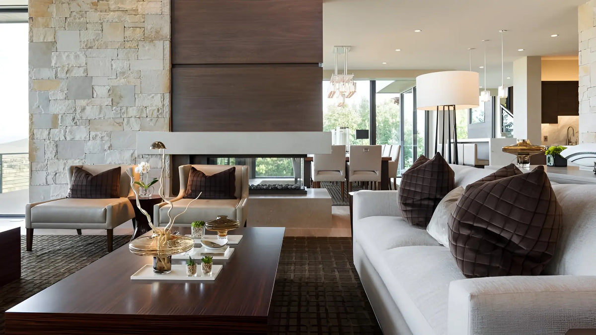 A modern living room interior featuring a white sofa with dark brown textured pillows, a dark wood coffee table, a stone wall, and an open-concept living space in the background.