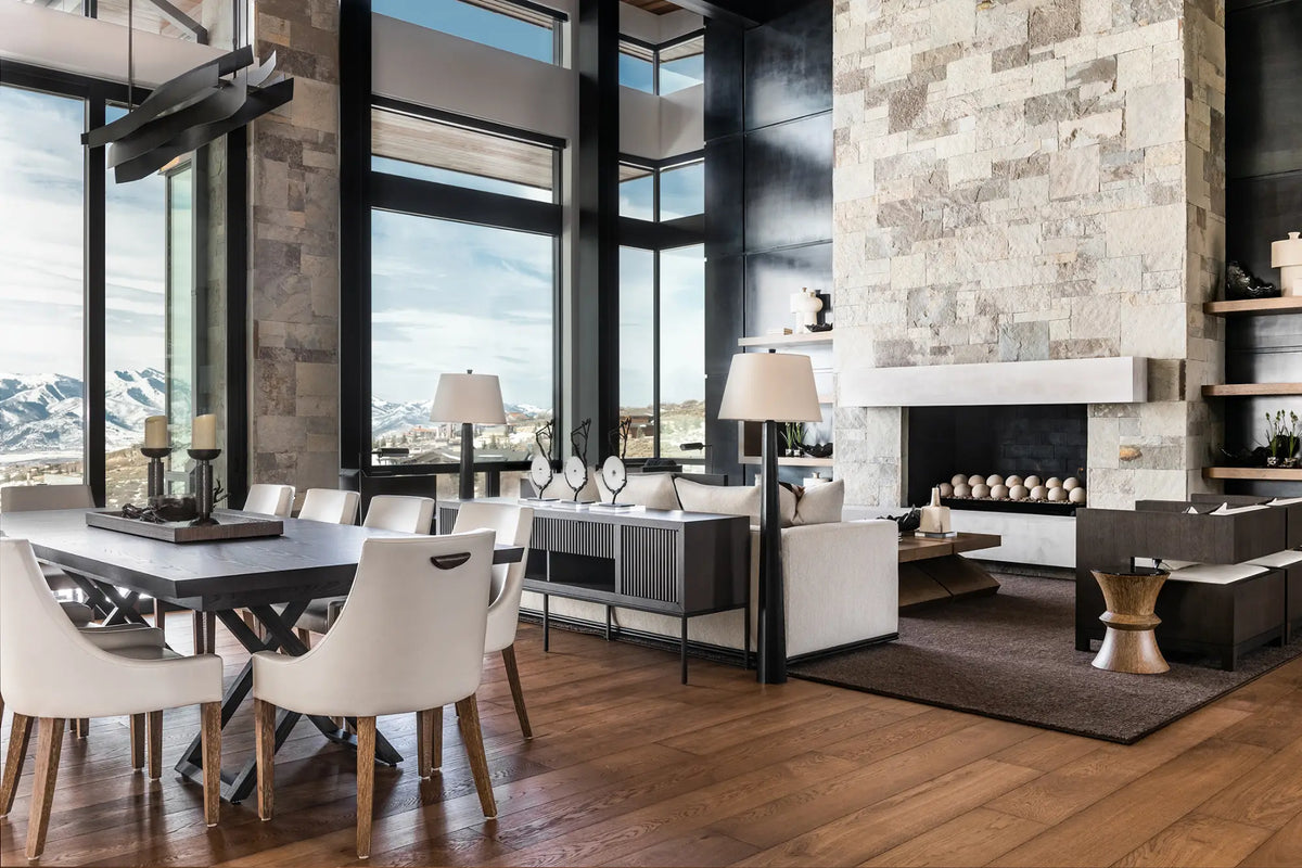 A modern dining room and living room interior with a dark wood dining table and white chairs, a stone fireplace, wood flooring, and large windows overlooking mountains.