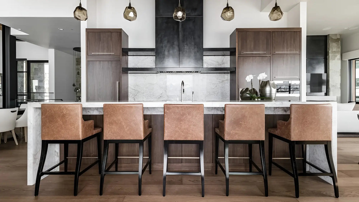Five modern bar stools with tan leather upholstery and black metal legs arranged along a marble-topped kitchen island in a contemporary kitchen setting.
