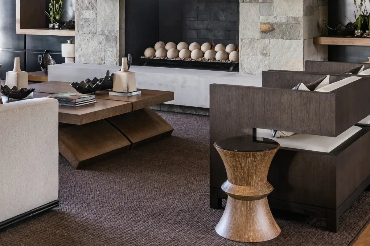 A modern living room interior featuring a dark wood coffee table, a dark brown sectional sofa with white cushions, a small round wooden side table, and a textured brown rug in front of a stone fireplace.