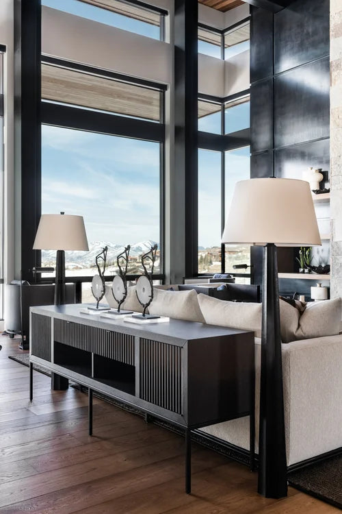 A modern living room interior featuring a dark wood and black console table with a ribbed front, flanked by two black floor lamps with cream drum shades. A light beige sofa is partially visible behind the table, set against a background of large windows with black frames and a high ceiling.