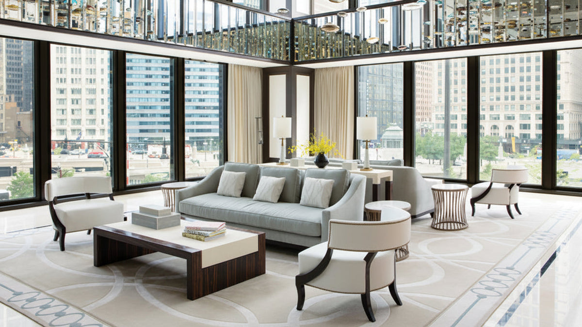 A modern living room with a light gray sofa, cream armchairs, a dark wood coffee table, and a large patterned rug, set against floor-to-ceiling windows with a city skyline view.