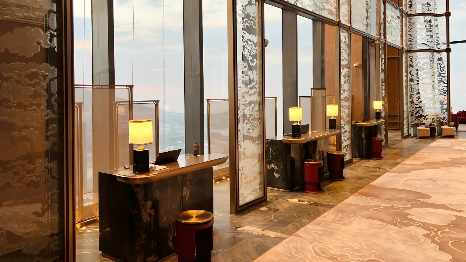 A luxury modern lobby interior with marble reception desks, lit table lamps, red and gold stools, and large floor-to-ceiling windows overlooking a city skyline.