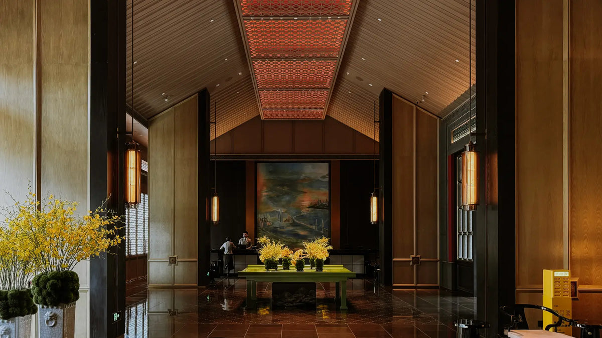 A grand, symmetrical interior lobby or hall with warm wood paneling, a patterned ceiling, polished dark floors, wall sconces, and a central table holding yellow flower arrangements in front of a large landscape painting.