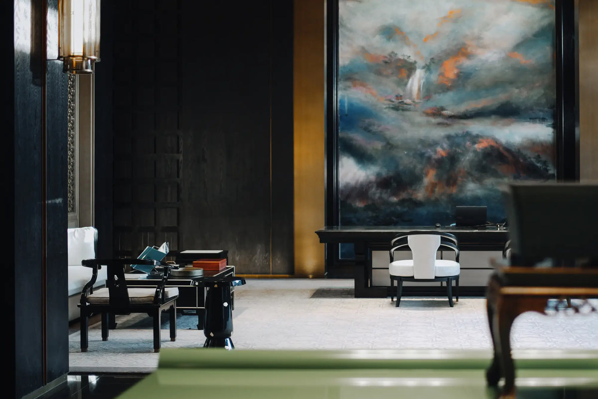 A large framed abstract landscape painting hangs on a dark wall in a modern, stylishly decorated interior with black furniture and warm lighting.