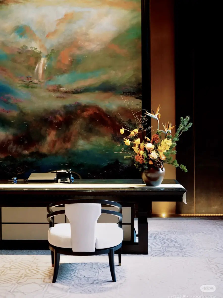 A large abstract landscape painting hangs on a dark wall above a long dark console table. In front is a white upholstered chair with a curved black frame, and a vase of mixed flowers sits on the table.