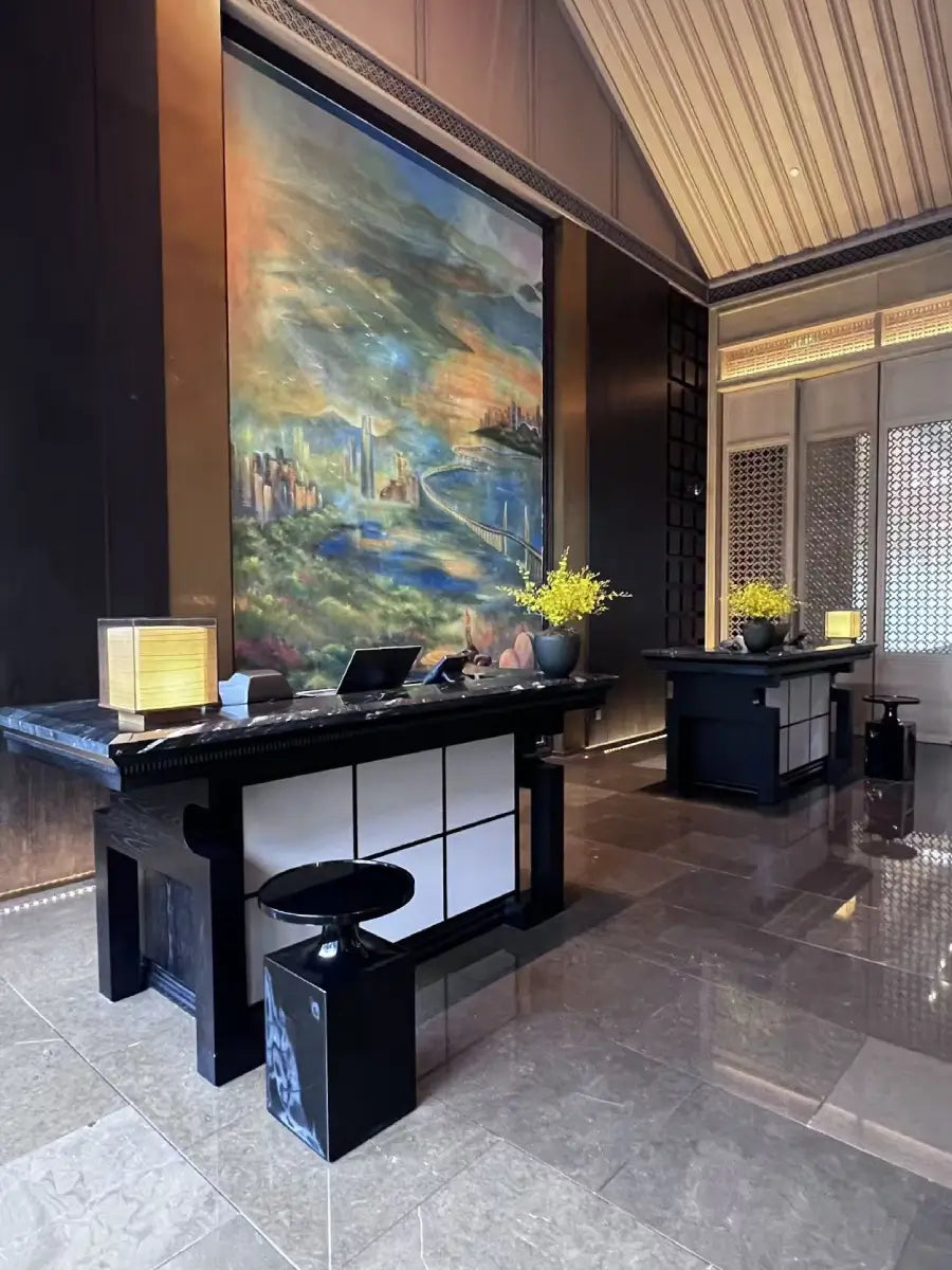 A modern reception desk area in a luxury hotel or lobby, featuring a large vertical landscape painting on the wall, black marble counters, white panel accents, and decorative yellow flowers.