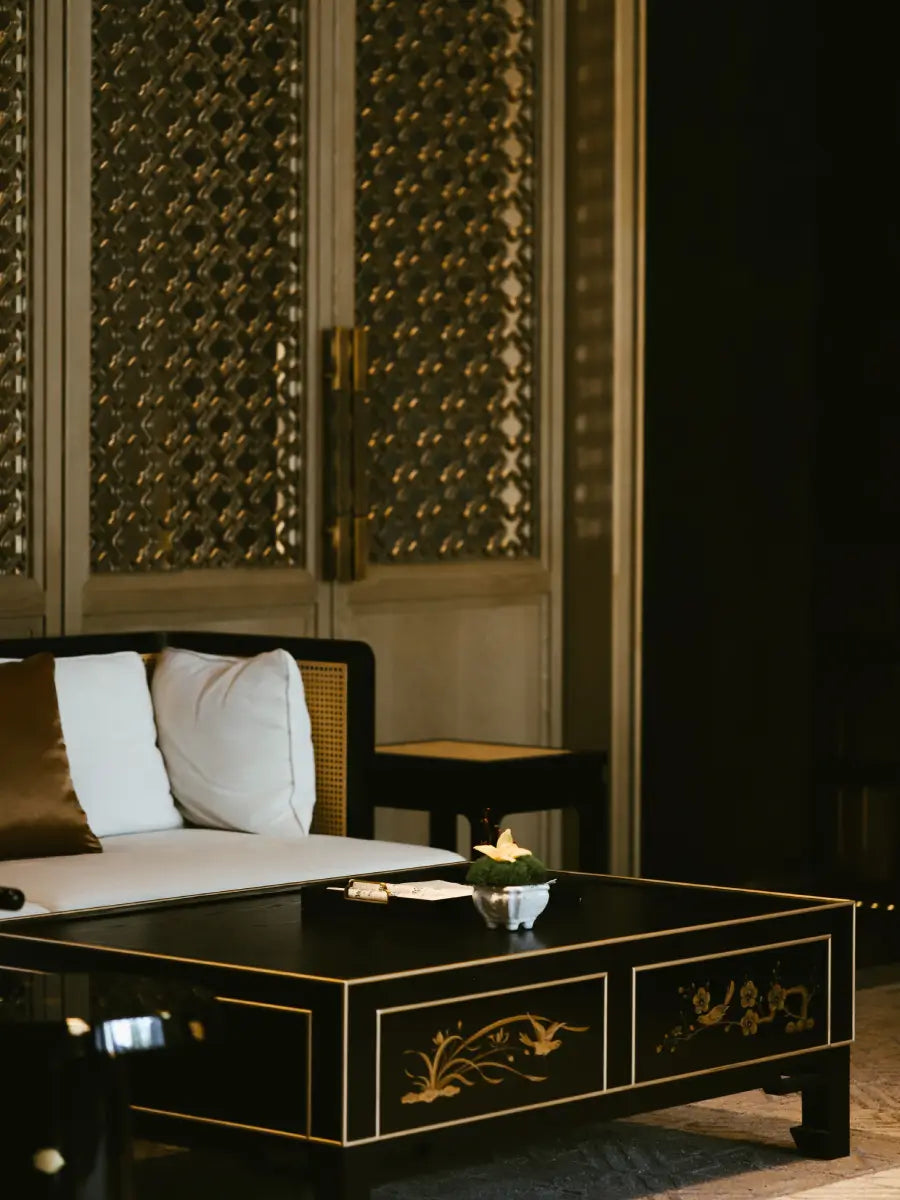 A black lacquered wooden coffee table with gold floral inlays, styled in a luxurious room setting with a low cushioned daybed and decorative screens in the background.