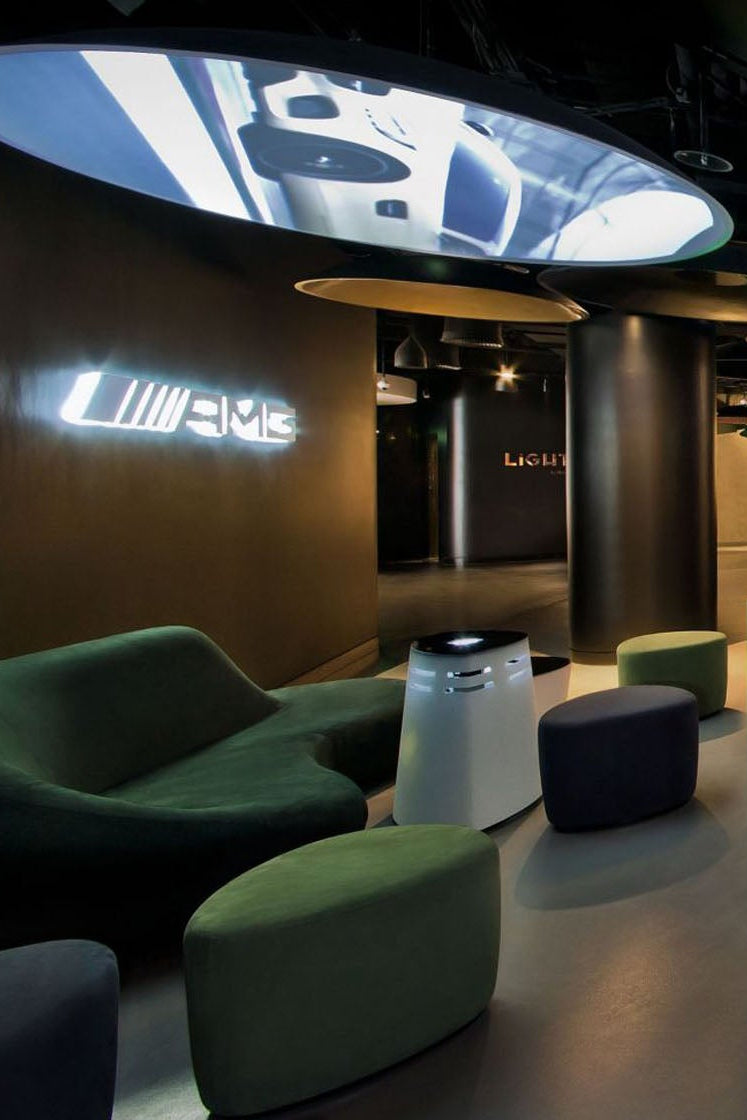 A modern interior lounge or showroom with dark curved seating, green and gray ottomans, large circular ceiling displays, and floor-to-ceiling windows overlooking a city night scene. A visible illuminated sign on the left wall reads LUMIERE, which appears to be the product brand.