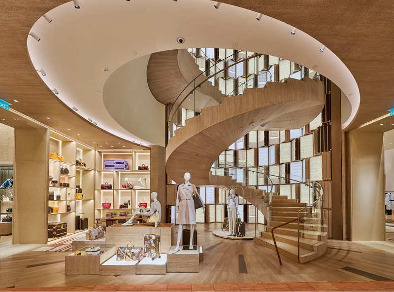 A luxury boutique interior featuring a prominent wooden spiral staircase, warm wood-toned ceilings, and mannequins displaying clothing on a raised platform.