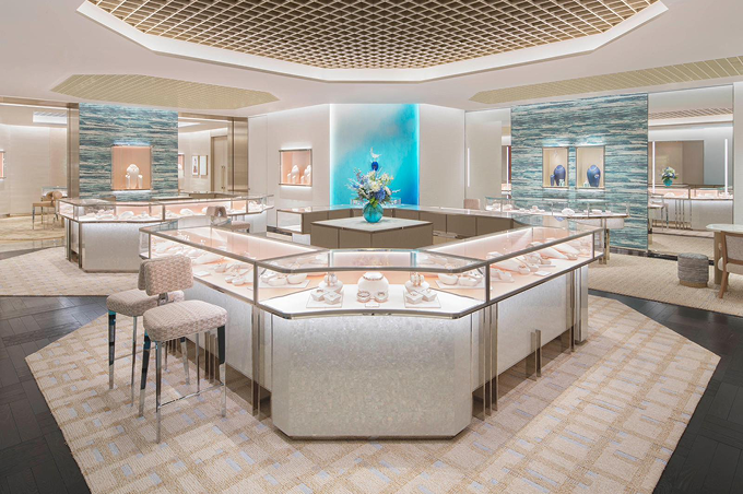 A luxury jewelry showroom interior with multiple glass display cases arranged in a U-shape and surrounding sections. The space features warm beige and cream tones, textured stone walls, a geometric ceiling, and a bright blue illuminated accent wall in the background.