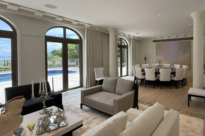 A luxury modern living room with a light beige two-seater sofa, a black leather sofa, a white sofa in the foreground, a dining area with a round dining table and chairs, arched windows looking out to a pool, and a large wall art piece.