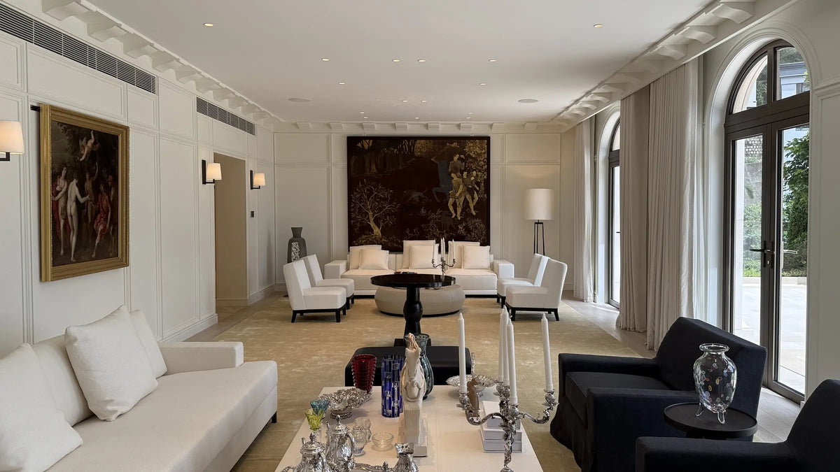 A luxury modern living room featuring a white leather sofa, white armchairs, a dark navy blue armchair, a round black coffee table, and a large framed artwork on the back wall.