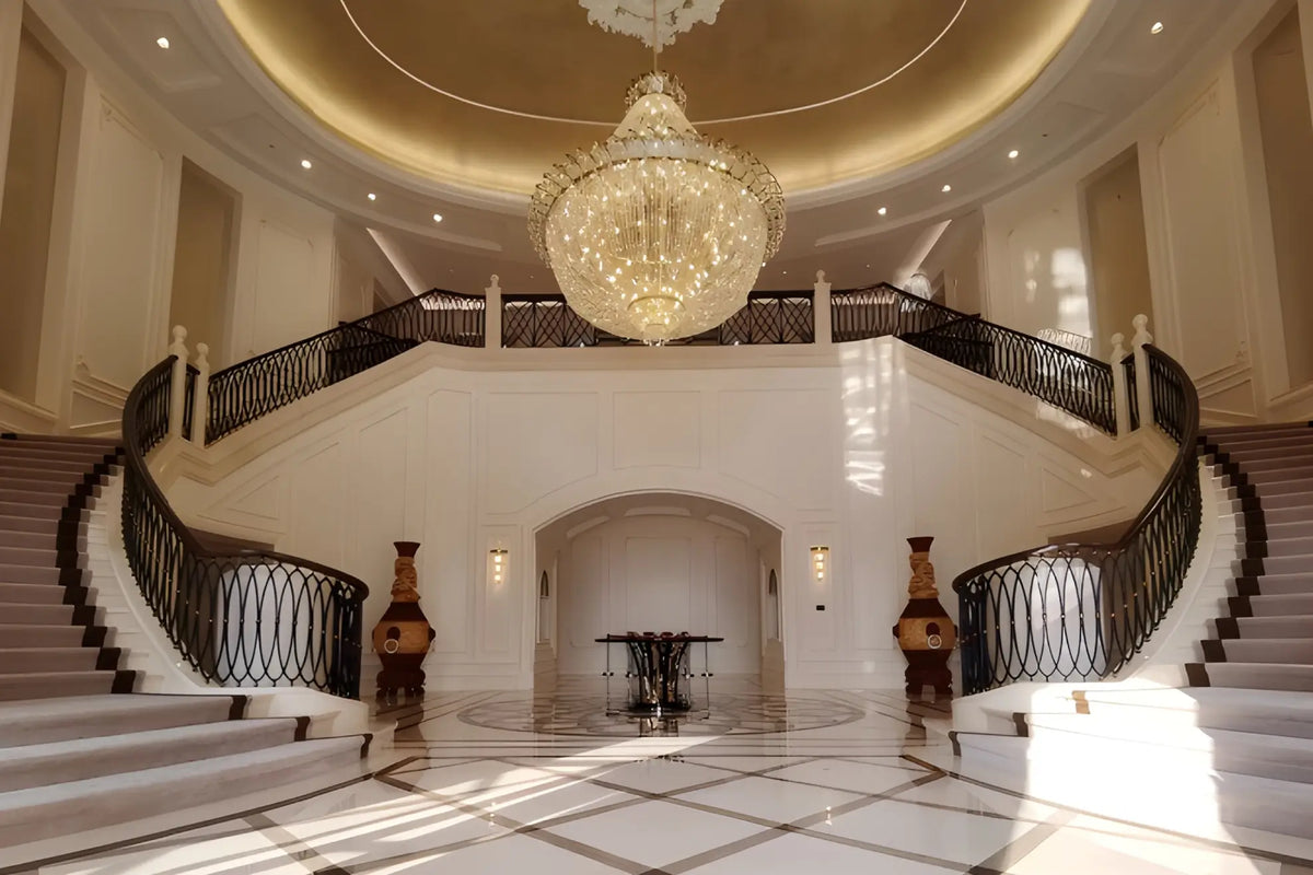 A grand interior staircase with sweeping curved steps, black metal railings, and a large ornate crystal chandelier hanging from a domed ceiling in a luxurious lobby.