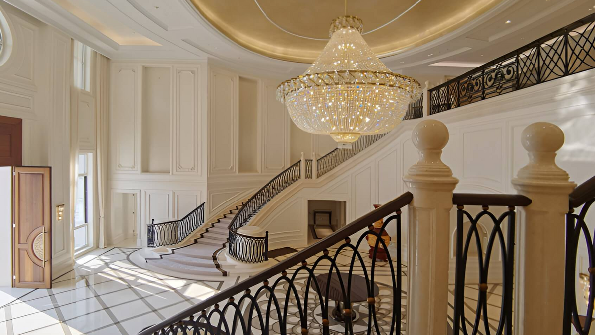 A grand luxury interior featuring a massive crystal chandelier hanging over a sweeping staircase with black wrought iron railings in a high-ceilinged elegant lobby.