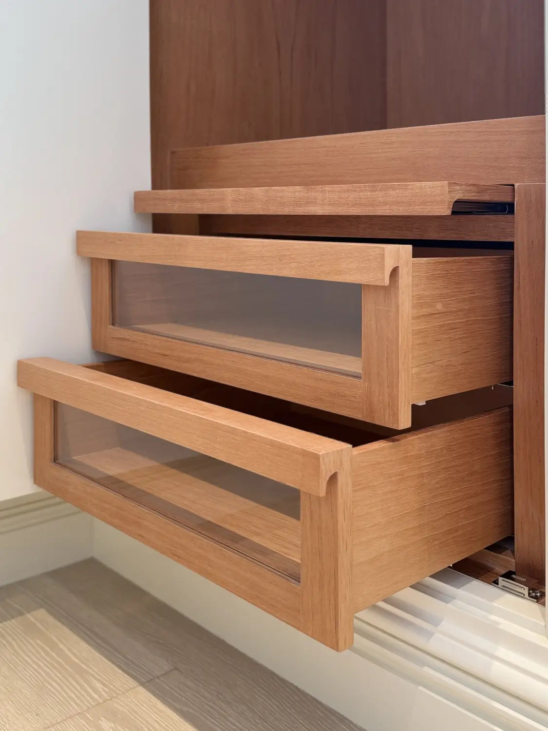 Close-up of three light oak wood drawers with glass-front panels, partially pulled out against a white wall and wood floor.