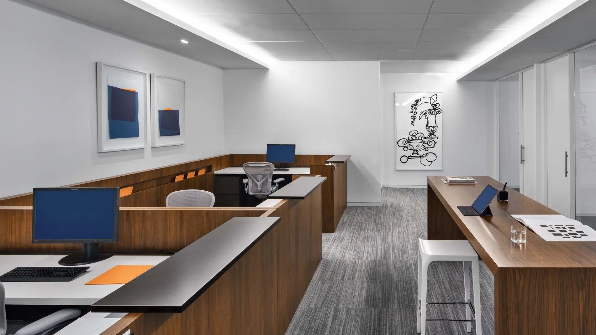 A modern office interior with wood partition desks, gray carpet, white walls, framed wall art, and a long reception desk with white stools.