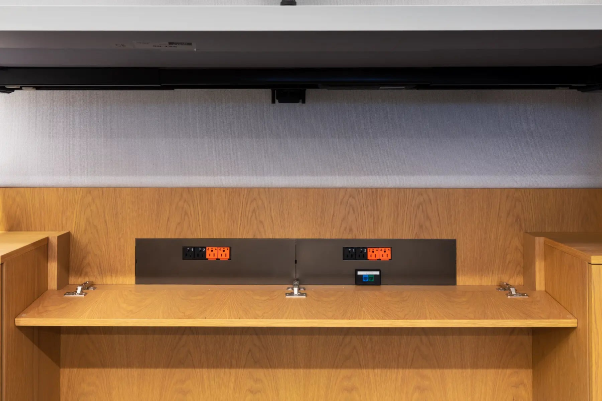 A close-up of a modern wooden interior with a built-in wood counter or shelf, a dark gray control panel with illuminated orange displays, and a light gray wall above.