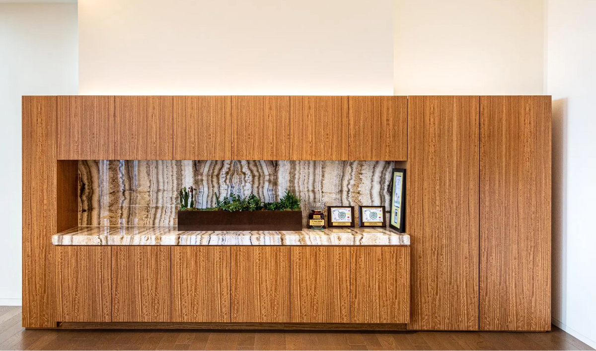 A modern wall-mounted wooden cabinet with a recessed center niche, featuring a polished marble countertop, a matching marble backsplash, and small decorative items including a planter and framed pictures.