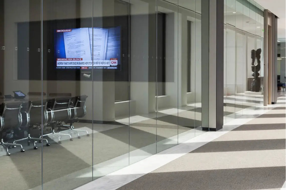 Modern office interior with clear glass walls and a meeting room visible through the glass, including a conference table, chairs, and a mounted display screen.