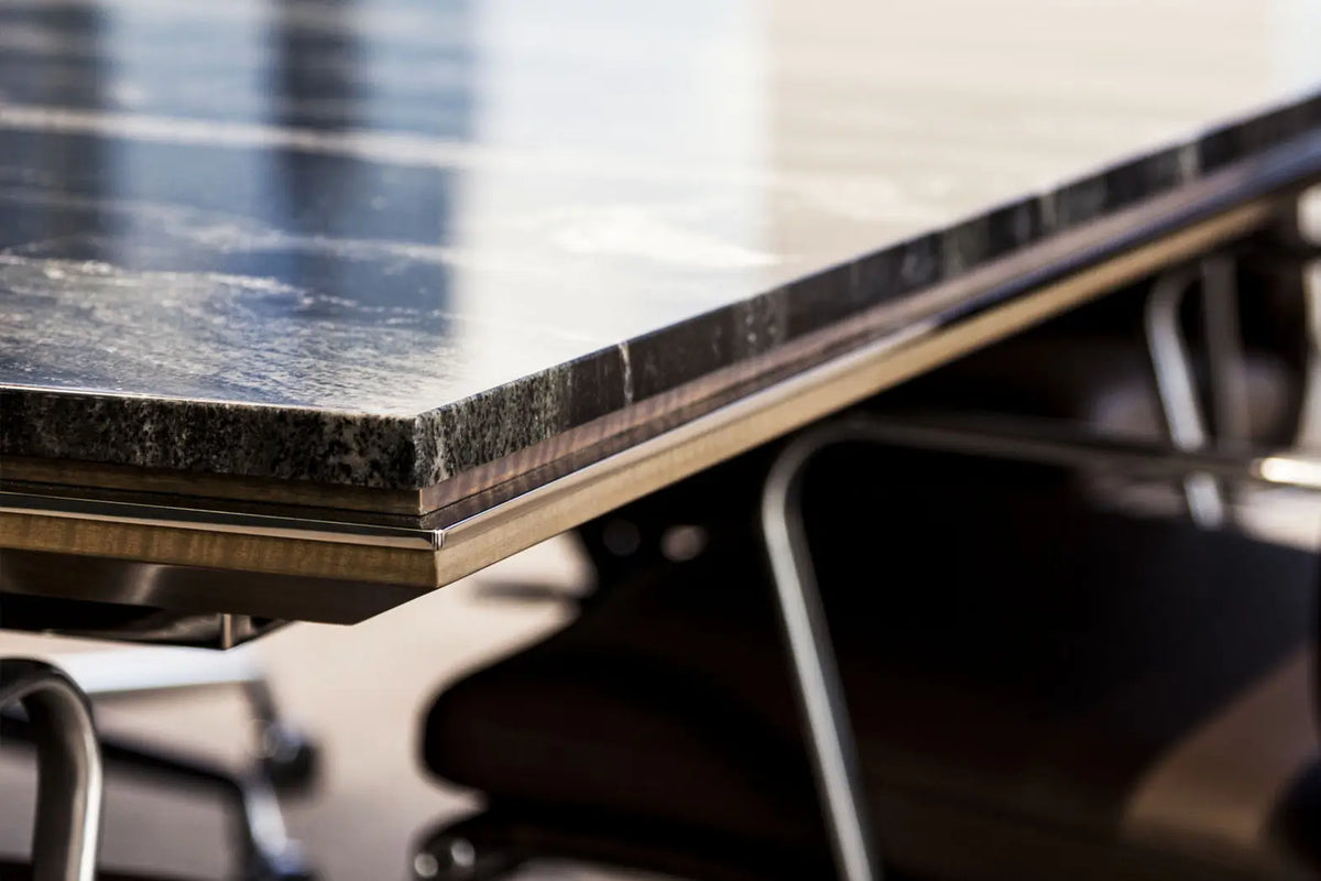 Close-up of a polished dark stone tabletop corner with a brushed metallic edge and blurred office chair legs underneath.