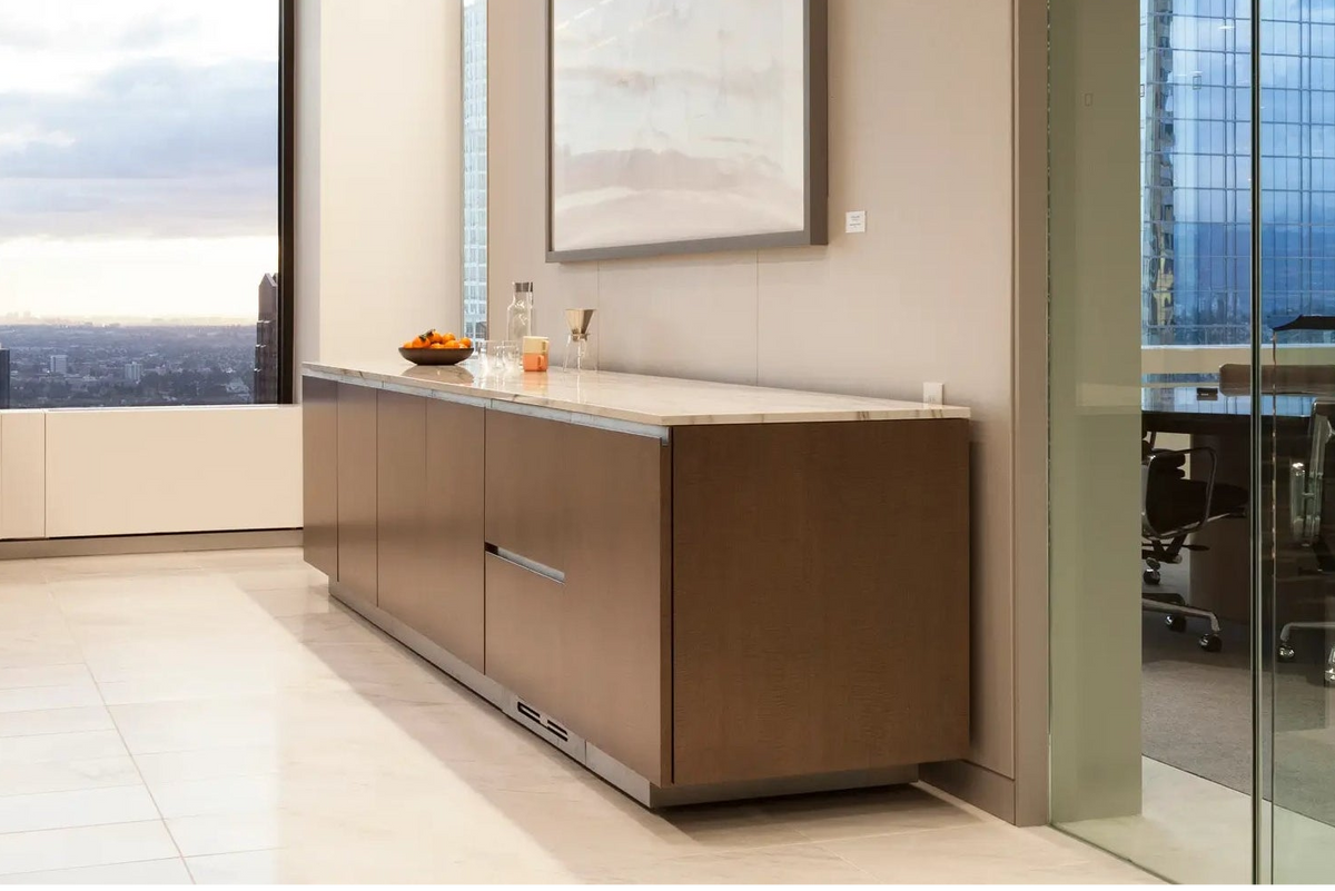 A modern office interior featuring a long brown wooden vanity cabinet with a white marble countertop, a large framed abstract print on the wall, and a glass-walled conference room in the background.