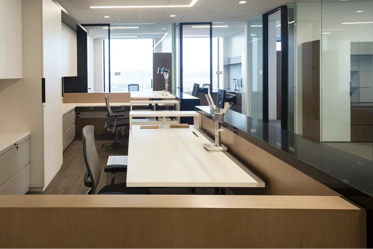 A modern office interior with long white desks, gray and black office chairs, glass partition walls, and large windows in the background.