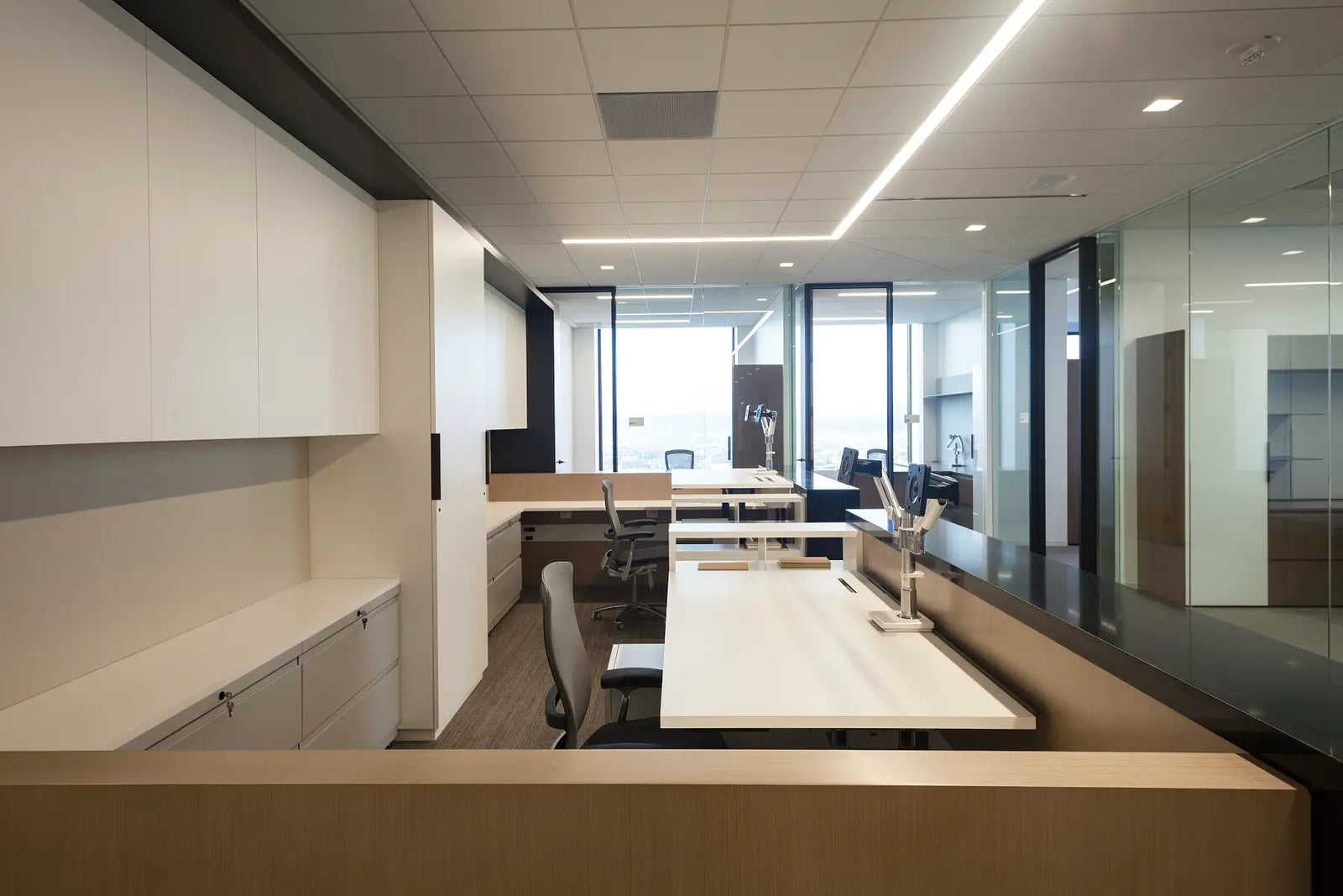 A modern office interior with long white desks, gray office chairs, glass partition walls, and a light wood counter in the foreground. The space is well-lit with overhead fluorescent lighting and recessed ceiling lights.