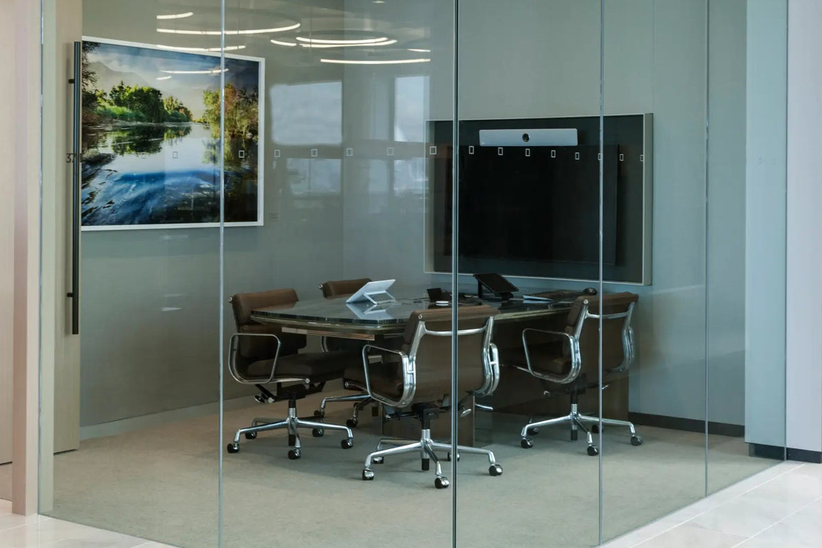 A modern glass-walled conference room with a round dark wood table, six black office chairs, a mounted screen, and a framed landscape picture on the wall.
